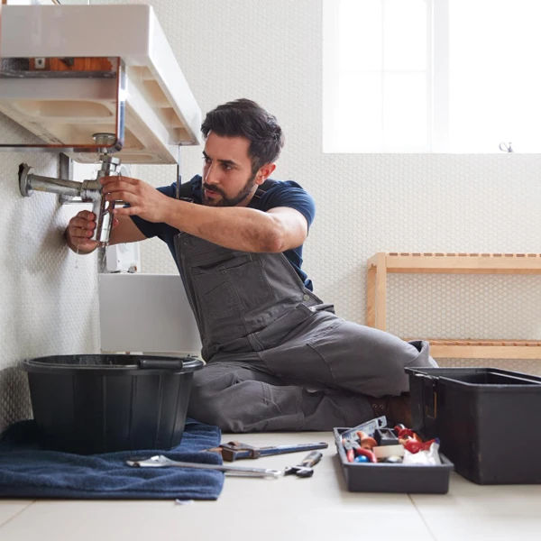 Encanador profissional de macacão cinza trabalhando no reparo do sifão sob uma pia de banheiro, com ferramentas e balde organizados ao lado.