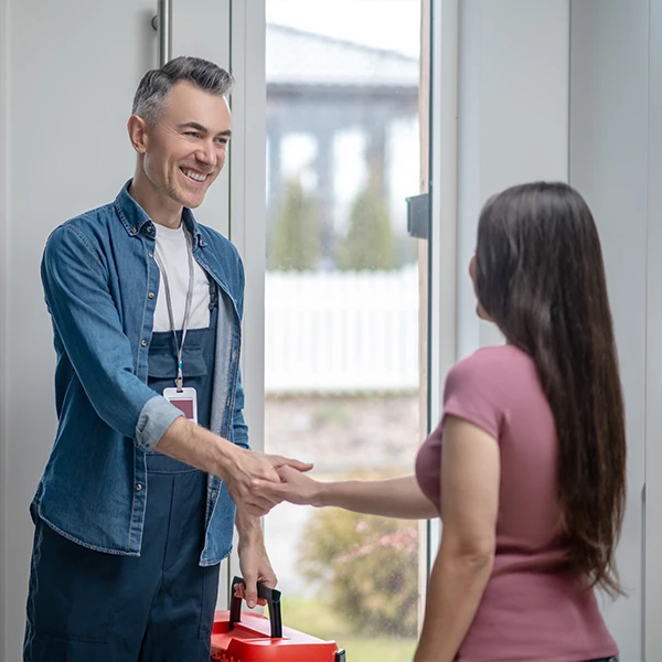 Um técnico de manutenção sorridente, vestindo uniforme azul e carregando uma maleta de ferramentas vermelha, cumprimenta uma cliente com um aperto de mão na entrada de uma residência.