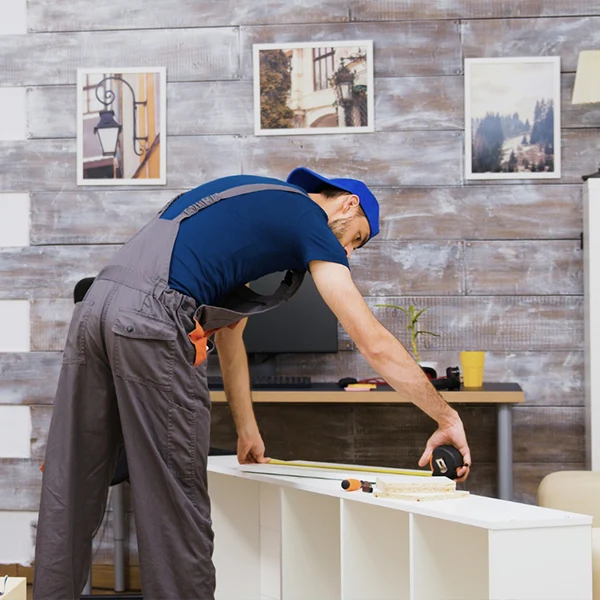 Homem de macacão cinza e boné azul, inclinado, usa uma trena métrica para medir a parte superior de uma prateleira branca inacabada, em um ambiente de escritório com parede de madeira.